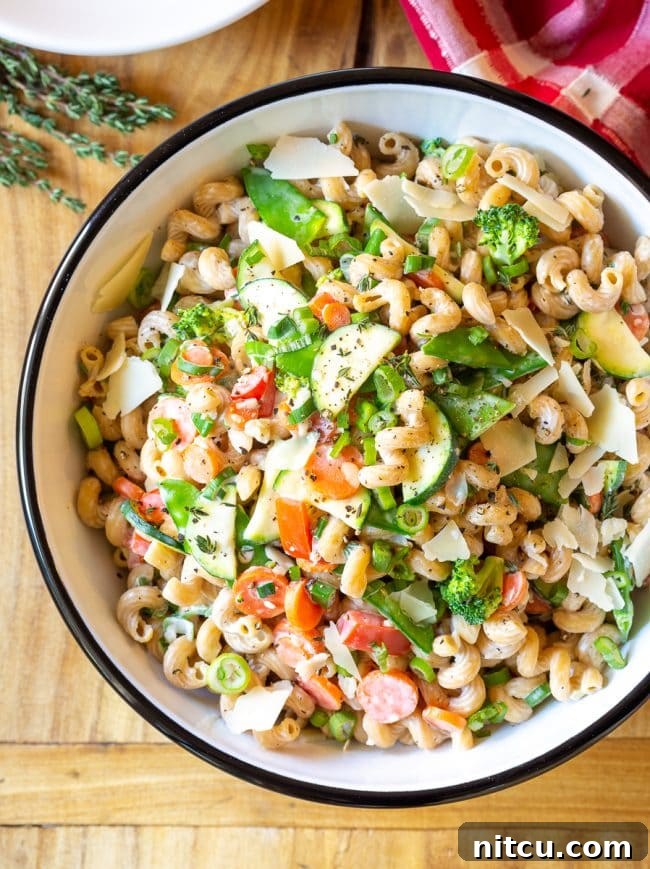 A top-down view of a serving bowl filled with creamy noodles and a colorful medley of chopped zucchini, carrots, broccoli, red peppers, and a garnish of shaved parmesan cheese.