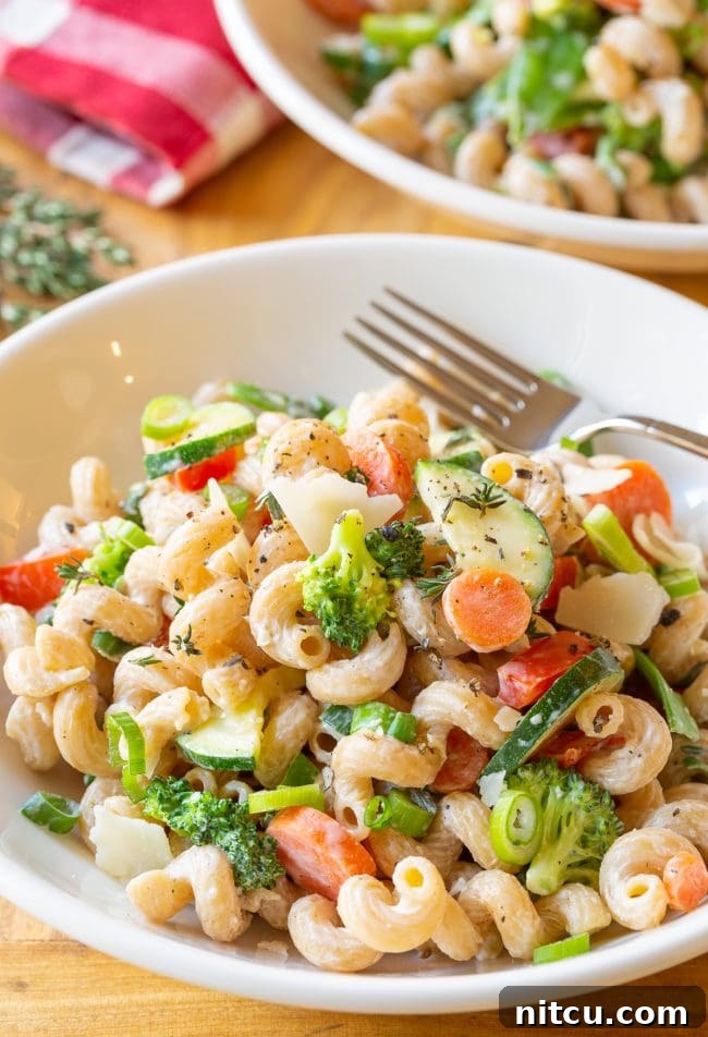 A white bowl of creamy noodles, generously mixed with chopped zucchini, carrots, broccoli, red peppers, and finished with shaved parmesan cheese.