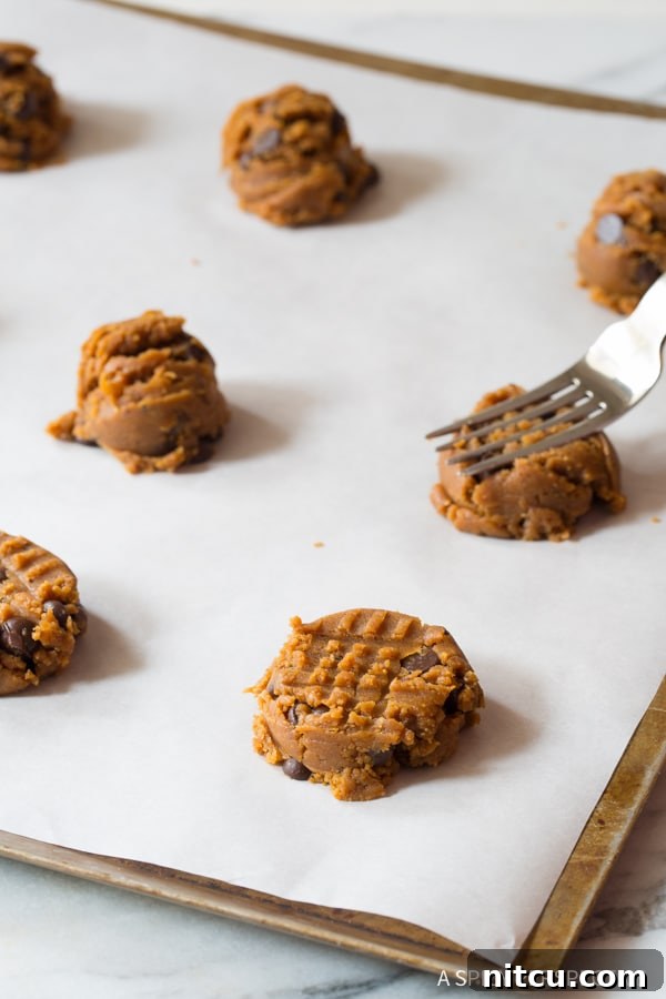 A baker using a fork to gently press down on individual cookie dough balls on a baking sheet, creating the classic crosshatch pattern.