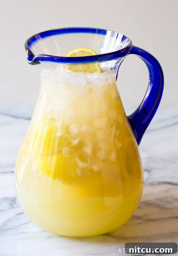 Side view of a clear glass pitcher brimming with vibrant Stevia Lemonade, artfully garnished with fresh lemon slices and a sprig of mint, resting on a rustic wooden surface.