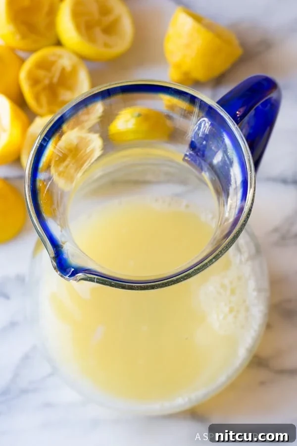 Freshly squeezed lemon juice being poured into a large, clear glass pitcher, ready for mixing.