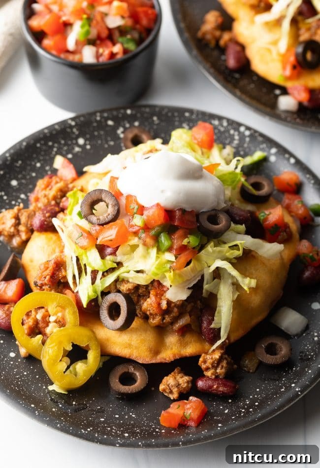 Navajo fry bread topped with seasoned ground beef, fresh lettuce, and other vibrant taco toppings on a rustic black plate, showcasing its golden crust and generous filling.