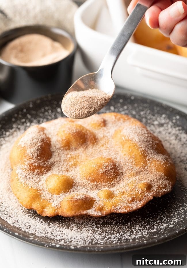 A hand gently sprinkling a generous amount of cinnamon sugar over a warm, freshly fried Indian Fry Bread, creating a sweet dessert.
