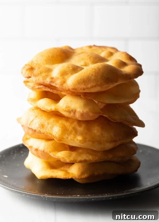A large stack of golden-brown Navajo fry bread, perfectly round and puffed, resting on a black plate, ready to be served or topped.