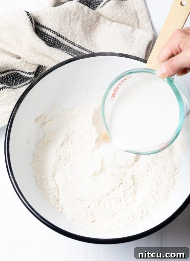 A hand carefully pouring hot liquid (milk and water mixture) into a bowl of dry ingredients, beginning the process of mixing the fry bread dough.