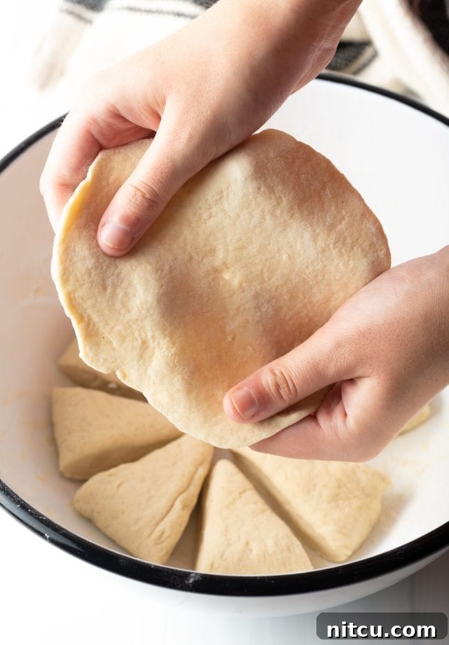A person gently stretching and pulling the edges of the soft dough by hand, shaping it into a thin, round tortilla-like disk, preparing it for frying.
