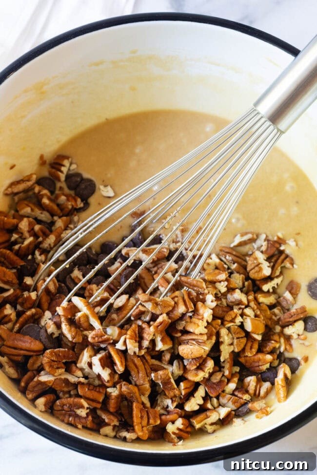 Close-up of a large mixing bowl with an eggy batter, as chopped nuts and dark chocolate chips are being whisked in.
