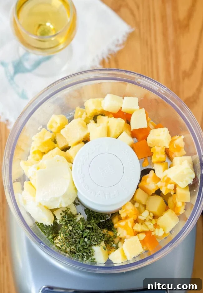 Overhead view of various cheeses and fresh herbs being added to a food processor, ready to be blended into a delicious spread.