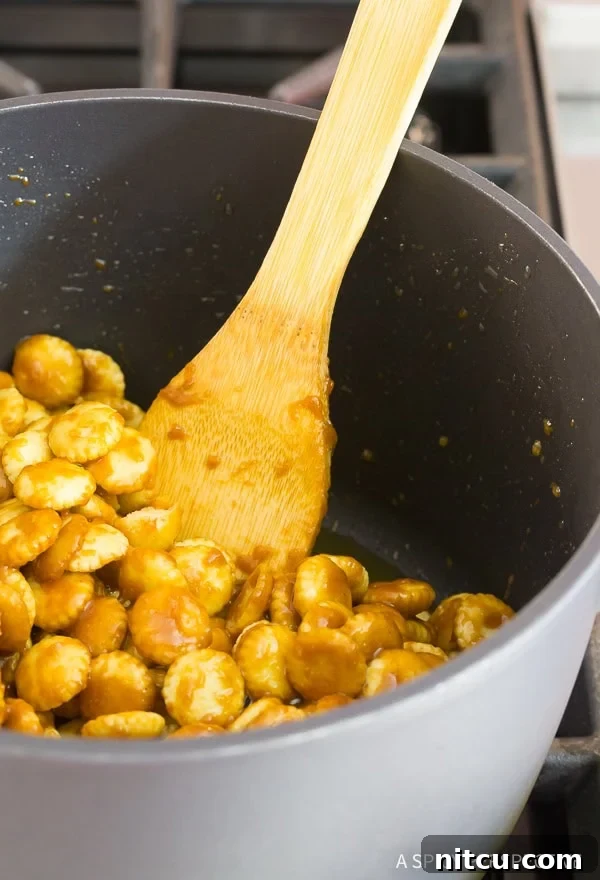A wooden spoon carefully coats oyster crackers with warm toffee mixture in a saucepan.