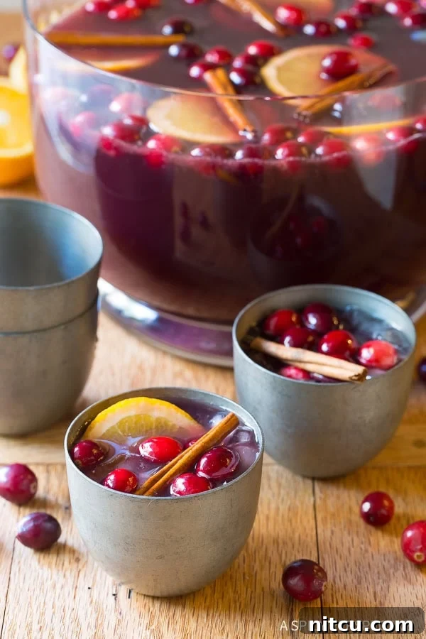 A beautiful punch bowl filled with Holiday Wine Punch, garnished with fresh cranberries and orange slices, with two elegant silver glasses ready for serving.