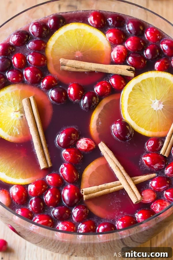 An overhead shot of a glistening punch bowl brimming with Holiday Wine Punch, adorned with vibrant cranberries, fragrant cinnamon sticks, and bright orange slices, ready for a festive gathering.