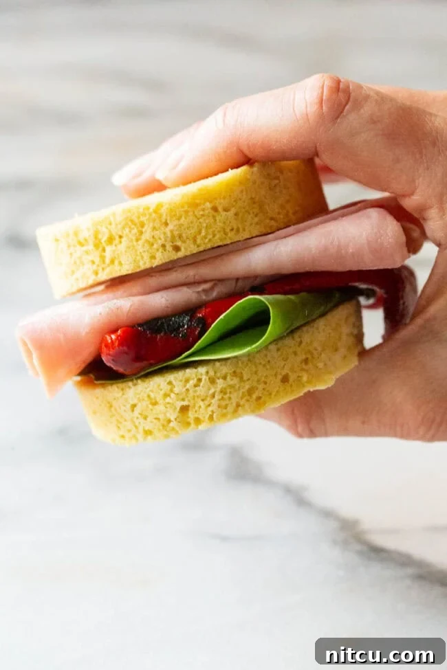 Hand holding a small sandwich made with two slices of low carb bread, sandwiching deli meat, roasted red pepper, and spinach leaves.