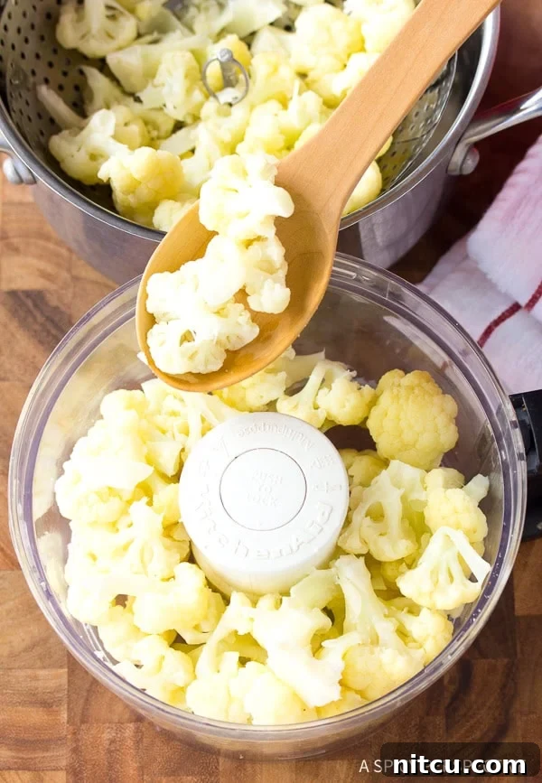 A wooden spoon carefully adding steamed and cooled cauliflower florets into a food processor for mincing.
