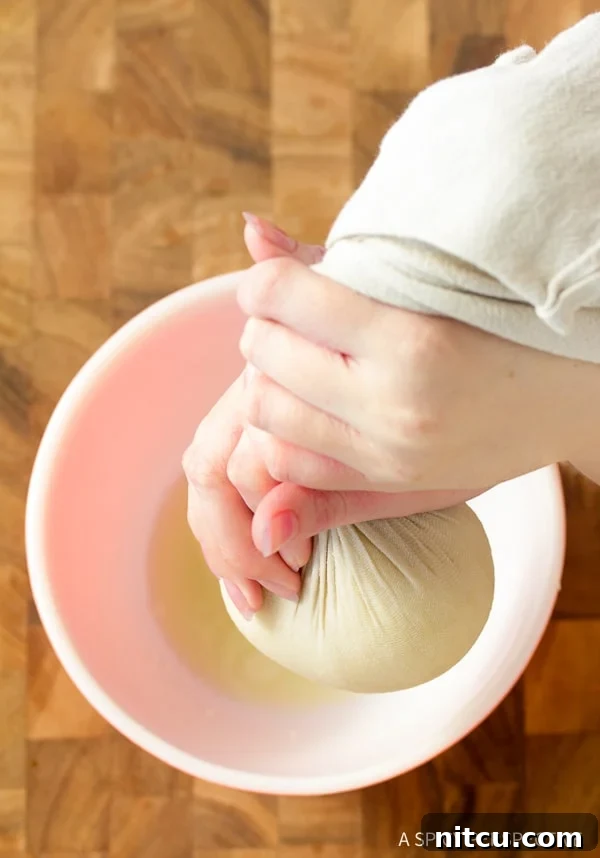 Hands firmly squeezing excess water from a bundle of steamed cauliflower wrapped in a cheesecloth over a bowl, illustrating a crucial step for crispy crackers.