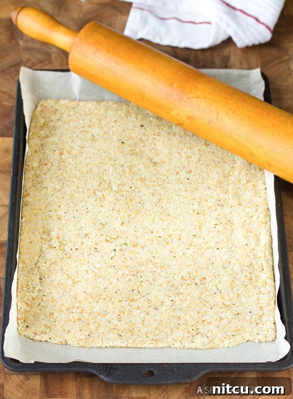 A baking sheet covered with a thin, evenly rolled layer of raw cauliflower cracker dough, ready for its first bake.