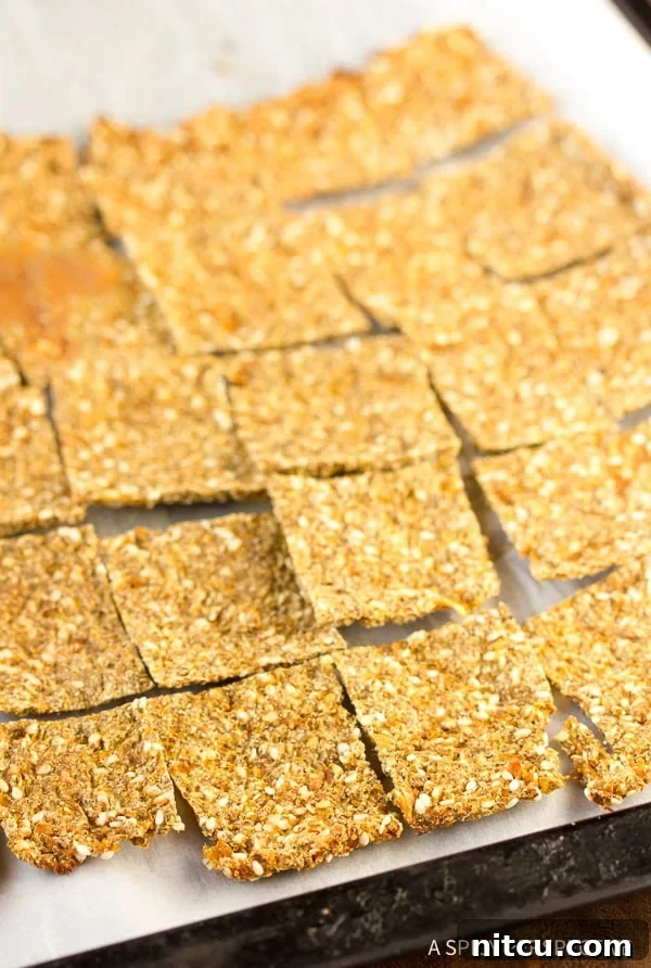 A close-up of a parchment paper lined baking sheet displaying an array of perfectly golden, square grain-free cauliflower crackers, highlighting their inviting texture.
