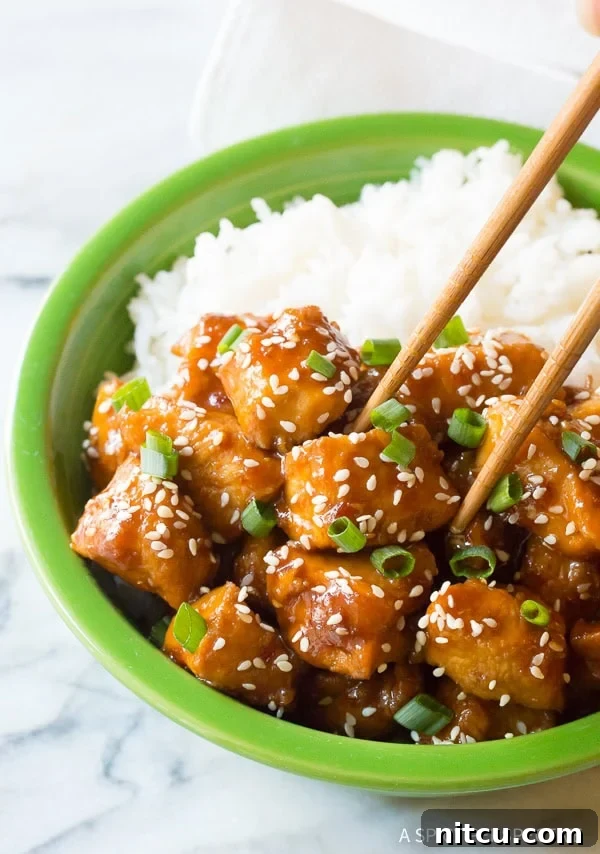 Chopsticks picking up a glistening piece of sesame chicken from a bowl of chicken and rice.