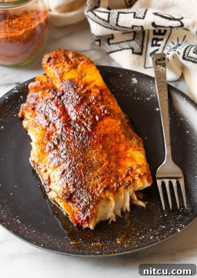 A perfectly cooked filet of fish, generously coated in vibrant blackened seasoning, rests on a sleek black plate alongside a metallic fork.