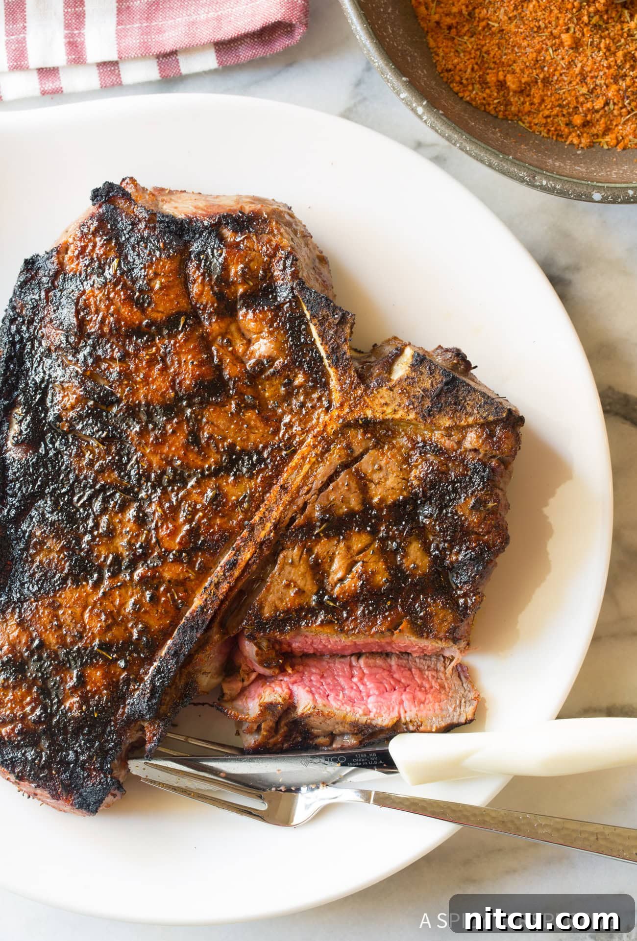A perfectly cooked steak, glistening with juices, rests on a white plate beside a fork and knife, ready to be enjoyed.
