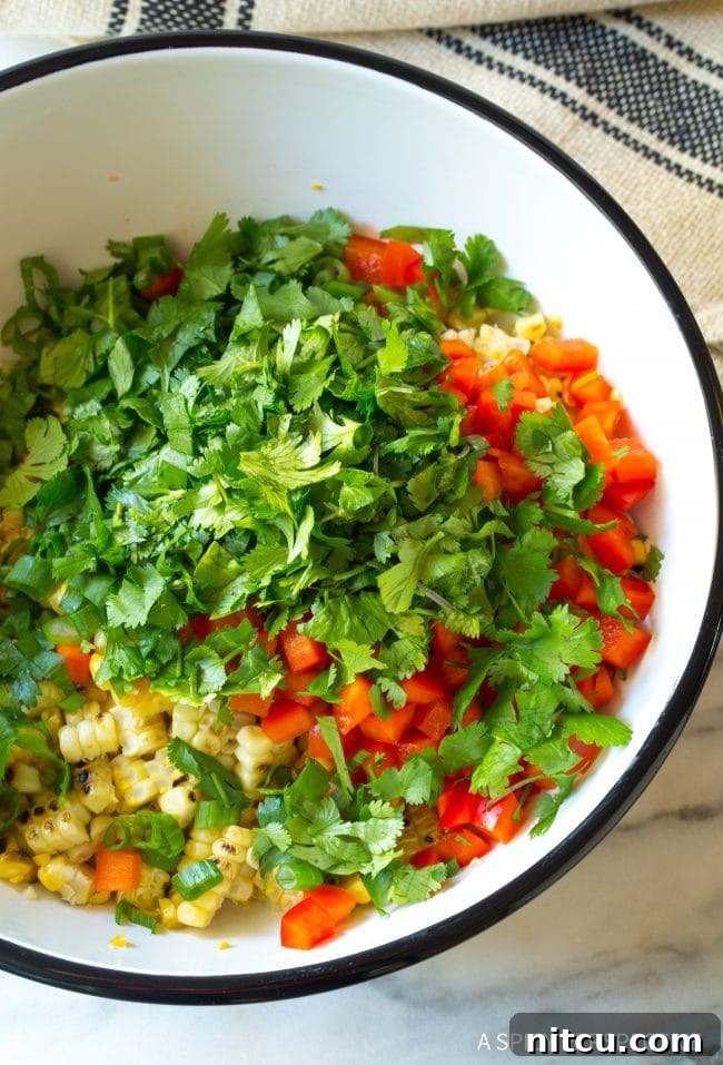 Overhead view of a bowl brimming with fresh, vibrant ingredients for street corn salad: colorful cilantro, red bell pepper, grilled corn, and green onions.