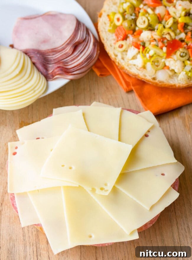 Swiss cheese slices being laid over the salami, completing the cheese and meat base for the New Orleans Muffuletta.
