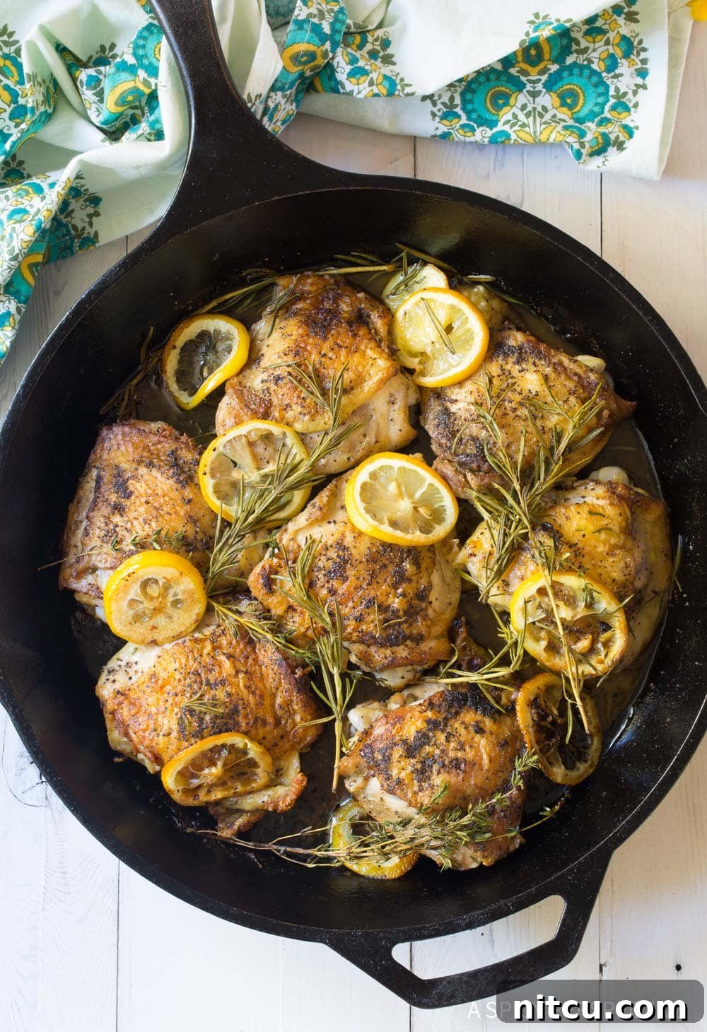 Oven Roasted Chicken Thighs in cast iron pan with lemon and herbs, showcasing golden skin and fresh ingredients.