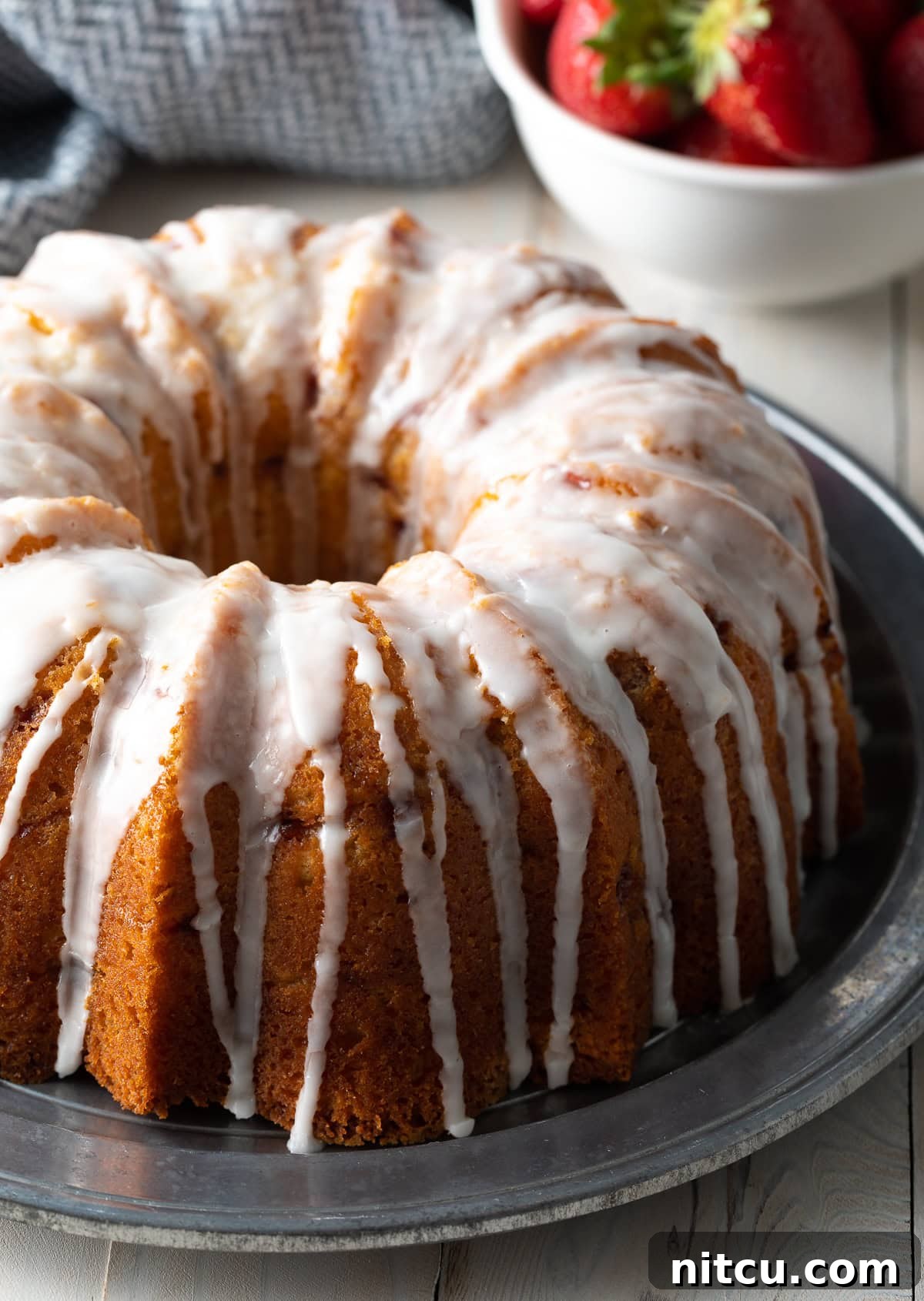 A beautifully glazed Strawberry Bundt Cake with lemon drizzle, resting on a elegant silver serving platter. The cake is adorned with fresh strawberry slices, highlighting its vibrant, natural ingredients.