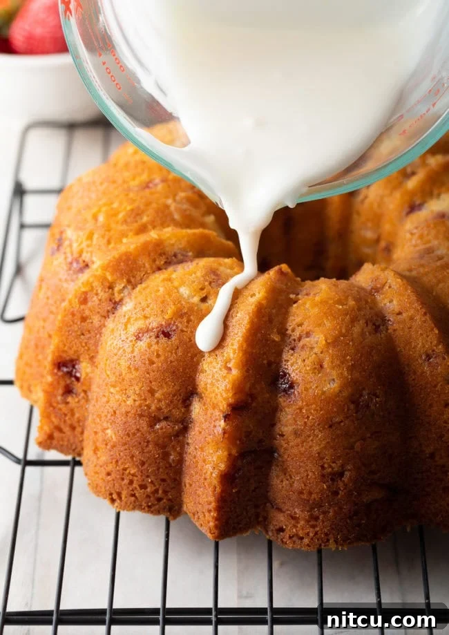 A freshly baked strawberry bundt cake being drizzled generously with a bright lemon glaze from a glass measuring cup.