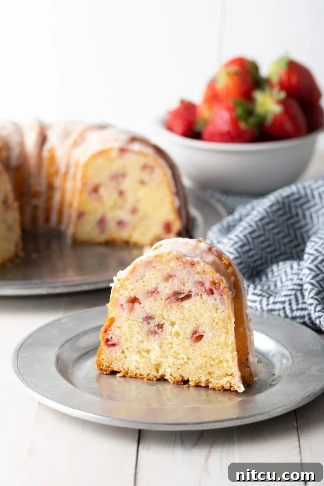 A close-up of a slice of strawberry bundt cake on a plate with the rest of the cake, beautifully glazed, in the background. The focus is on the inviting texture of the cake.
