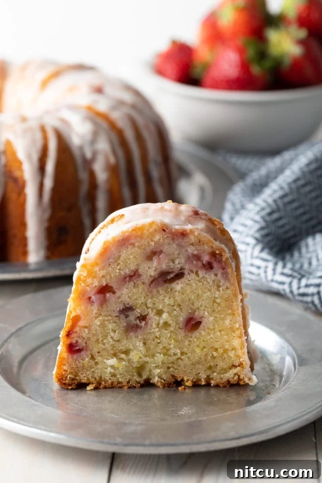 A perfect slice of the Strawberry Bundt Cake, showcasing its moist interior, studded with fresh strawberries, and topped with a luscious lemon glaze. The slice sits on a silver plate, ready to be enjoyed.