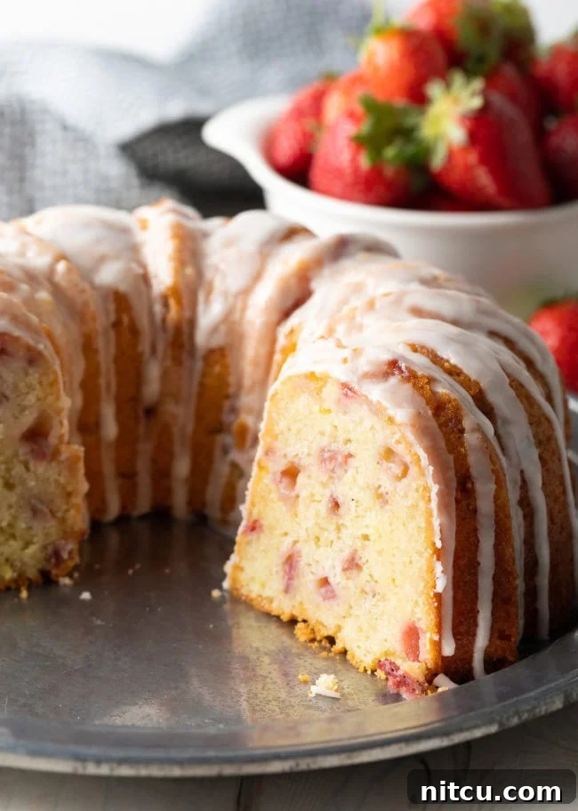 A freshly baked Strawberry Bundt Cake with a slice removed, revealing the soft, moist interior dotted with cooked strawberries. The cake is lightly glazed and sitting on a wire rack to cool.