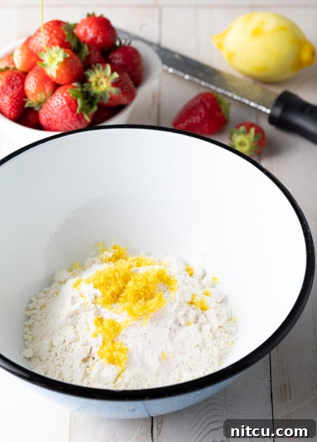 A mixing bowl filled with sifted flour and lemon zest, with a separate bowl of fresh strawberries beside it on a kitchen counter, preparing for the cake batter.