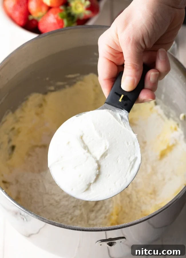 Strawberry cake batter in a mixing bowl with a hand holding a measuring cup with yogurt above it, illustrating the addition of wet ingredients.