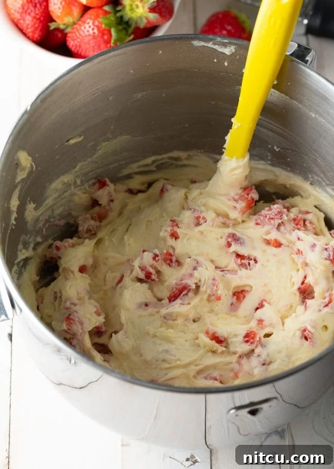 Strawberry cake batter with strawberries added and being gently stirred with a yellow spatula, ensuring even distribution.