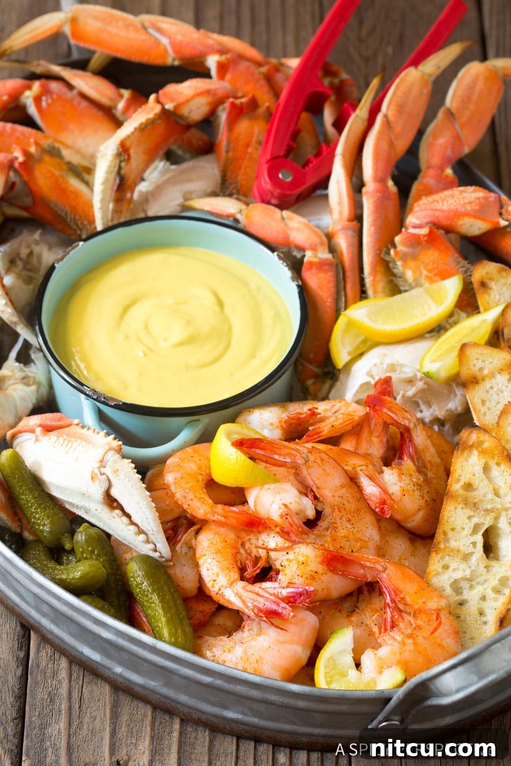 A vibrant seafood platter featuring cooked shrimp, crab legs, and lemon wedges, with a small bowl of spicy mustard seafood sauce at the center.