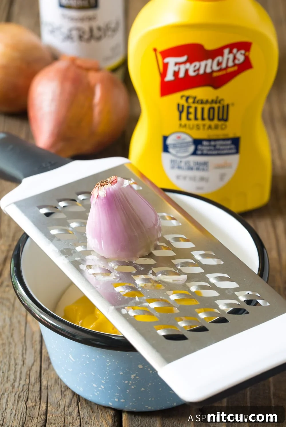 A hand grating a shallot directly into a bowl of the mustard sauce mixture, ensuring fine pieces.