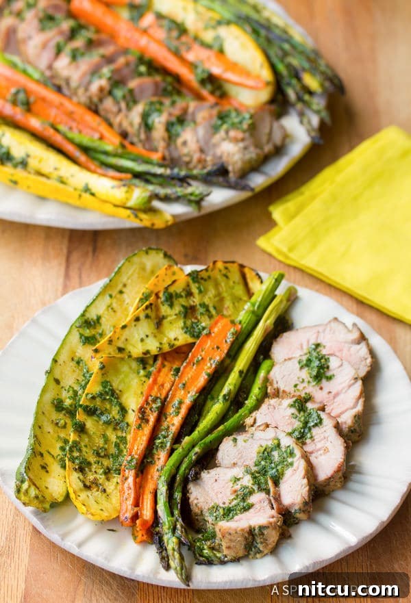 Sliced grilled pork tenderloin with chimichurri and roasted vegetables on a plate, garnished with fresh herbs, ready to be served.