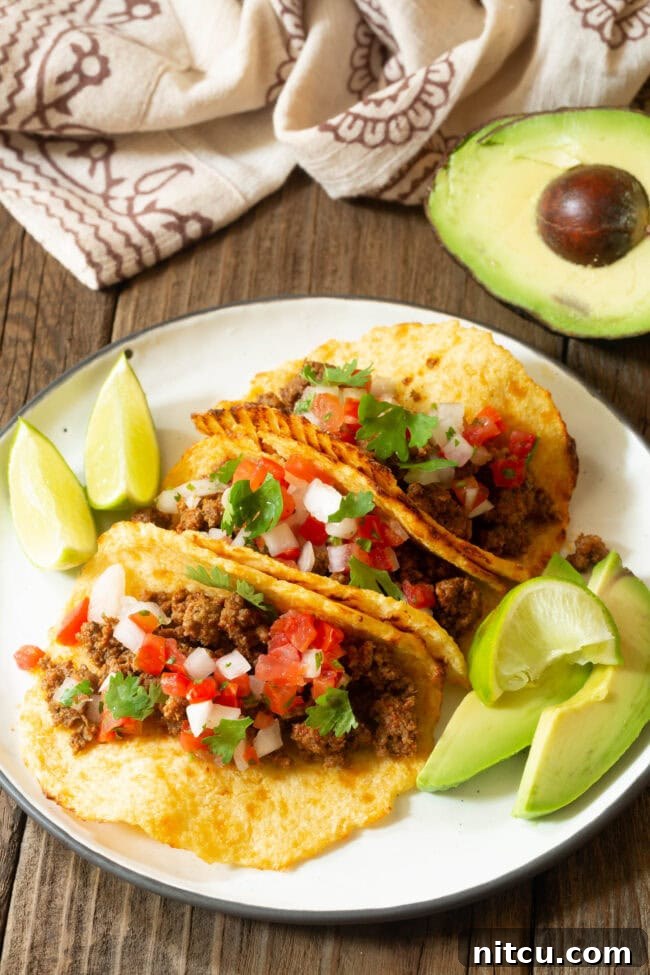 Three perfectly assembled tacos on a plate with lime wedges and avocado slices on the side.