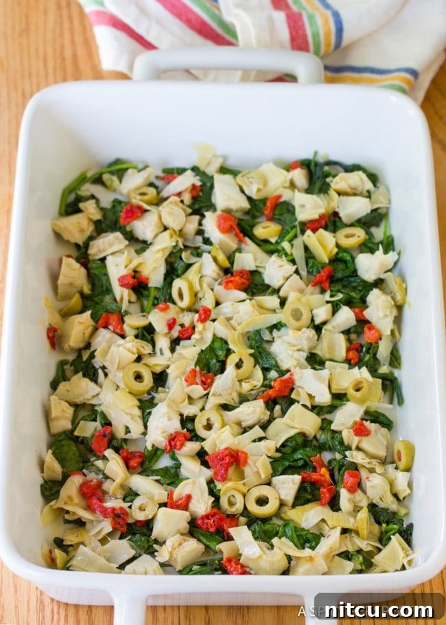 A casserole dish with vibrant green spinach and tangy artichoke salad evenly layered, awaiting the egg mixture.