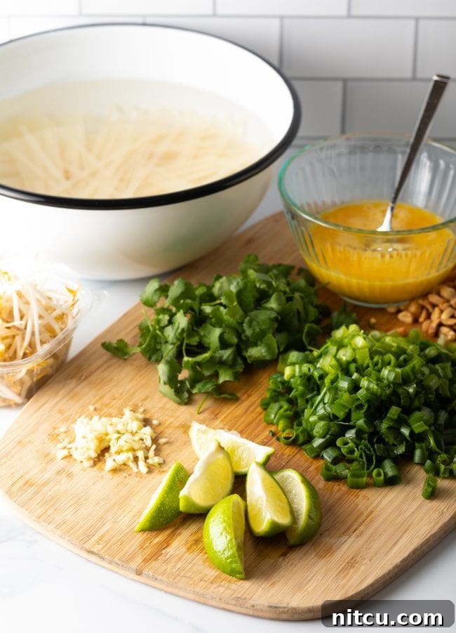 A wooden cutting board displaying neatly arranged Pad Thai ingredients: minced garlic, fresh lime wedges, chopped green onions, cilantro, and a bowl of beaten eggs.
