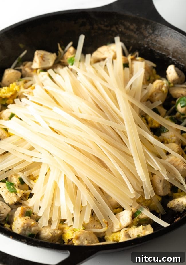 Adding cooked rice noodles on top of the vibrant stir-fry mixture of scrambled eggs, bean sprouts, green onions, garlic, and chicken in a large skillet.