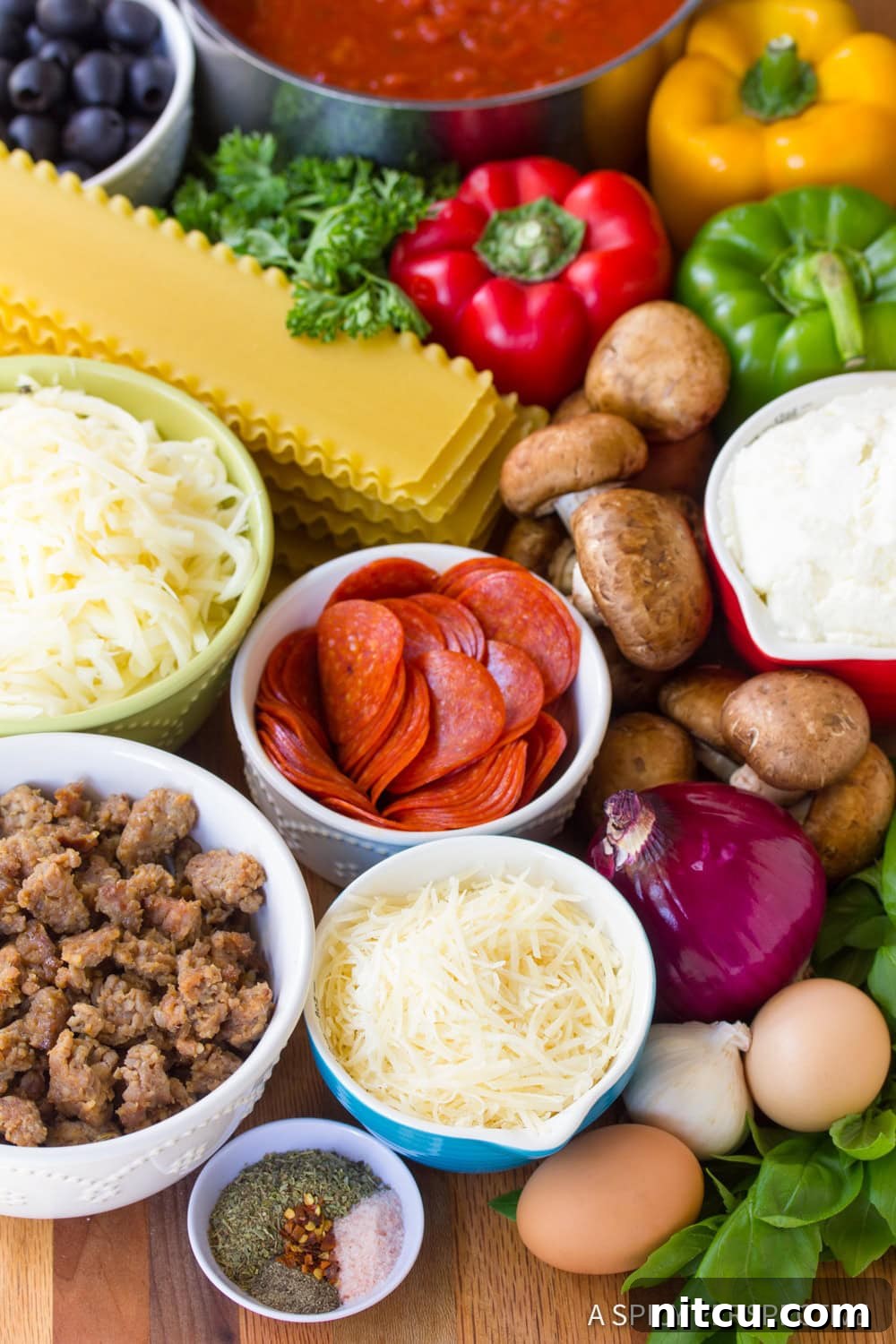 A vibrant display of fresh ingredients for Supreme Pizza Lasagna, including uncooked lasagna noodles, mushrooms, pepperoni, colorful bell peppers, shredded mozzarella and Parmesan cheeses, an onion, eggs, Italian sausage, and various spices.