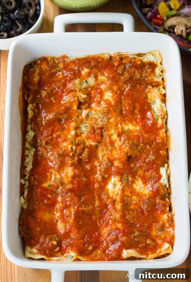 An overhead view of the Supreme Pizza Lasagna being carefully layered in a white baking dish, showing the organized placement of noodles, cheese, sauce, and vegetables.