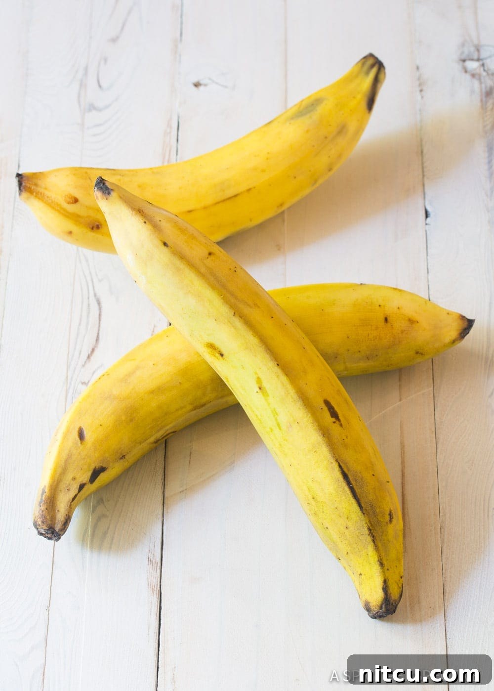 Three ripe yellow plantains with black spots resting on a kitchen counter, ready for preparation.