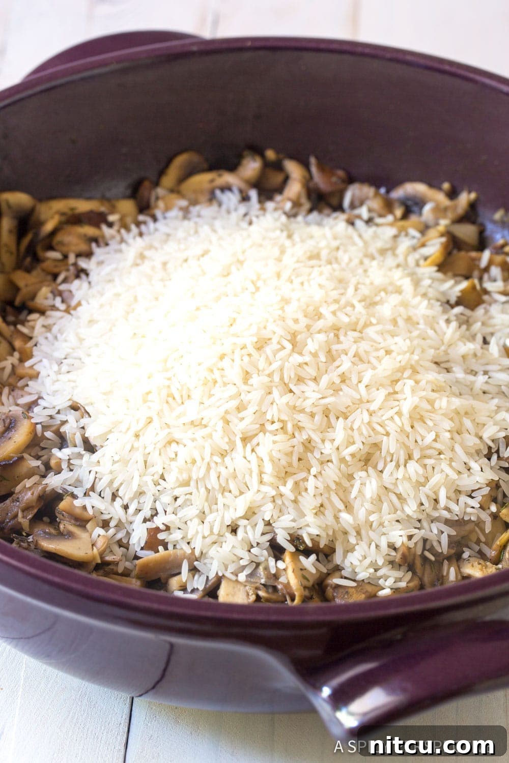 A top-down view of uncooked long grain rice resting on a bed of savory sautéed mushrooms in a pan, ready for the liquid to be added.