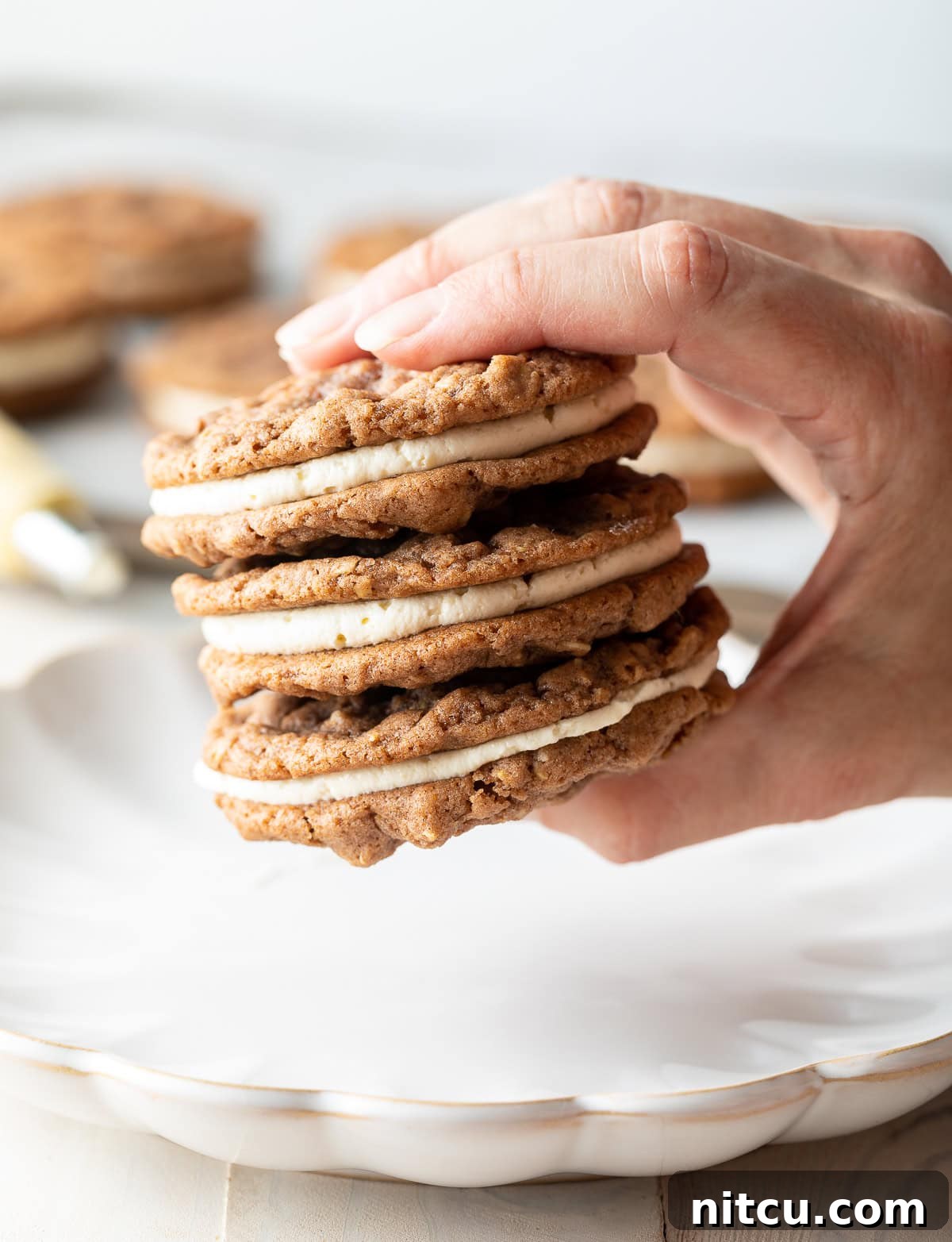 A hand holding three perfectly assembled Homemade Oatmeal Cream Pies above a serving tray.