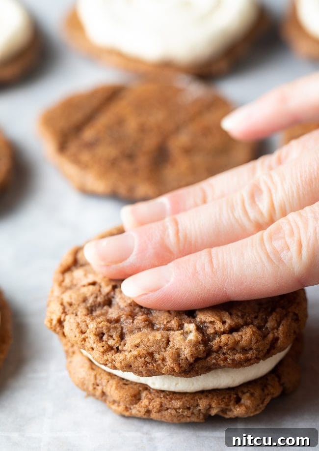 A hand gently pressing the top layer of an oatmeal cookie over the cream filling to complete the pie.