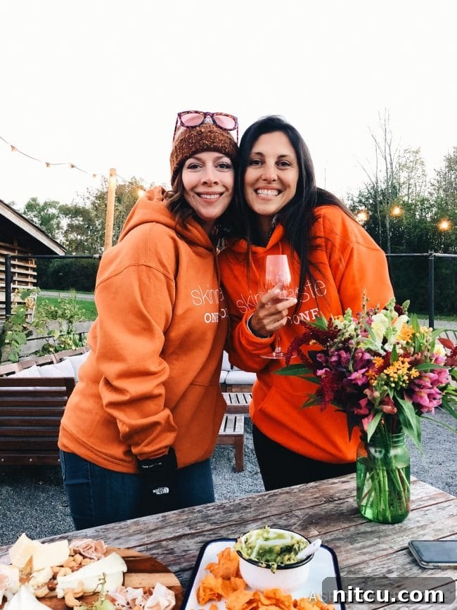 Two cheerful women, both sporting orange hoodies emblazoned with 'SkinnyTaste,' one raising a wine glass in celebration.