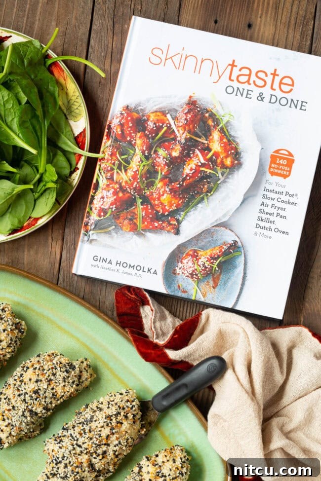 The SkinnyTaste One & Done Cookbook resting open on a rustic wooden table, alongside a plate of freshly cooked chicken roll-ups and a vibrant bowl of fresh spinach.