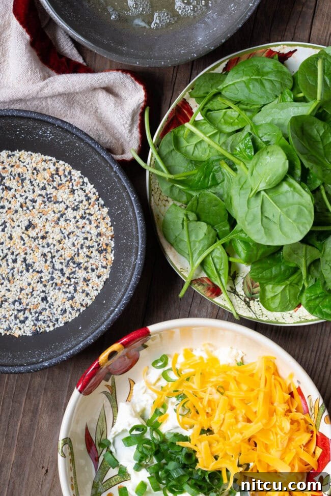 A vibrant display of recipe ingredients, featuring a fresh bowl of baby spinach, a shallow dish brimming with aromatic everything bagel seasoning, and a bowl containing a perfectly combined mixture of cream cheese, freshly sliced green onions, and shredded cheddar cheese.